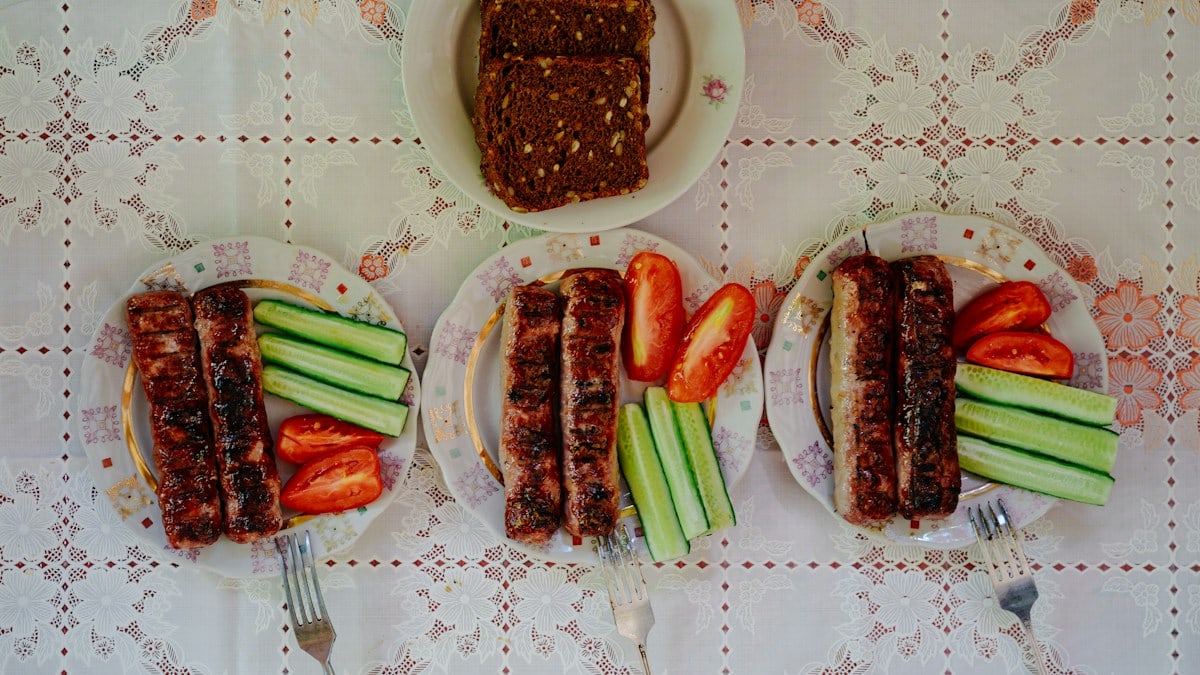 Three plates of grilled sausages with sliced cucumbers and tomatoes beside dark rye bread